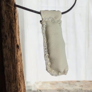 May include: A white leather pouch with a decorative stitch pattern hangs from a black cord. The pouch is made of soft, supple leather and has a smooth, even texture.