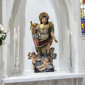 May include: A white statue of an angel with wings, holding a sword and standing on a base with three cherubs. The angel is wearing a gold crown and a colourful robe. The statue is in a white alcove with white candles on either side.
