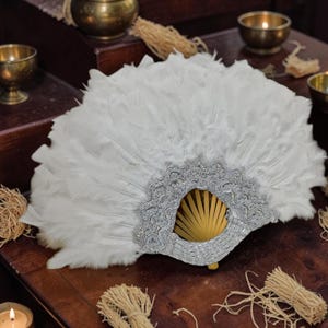 May include: A handheld folding fan featuring dense white feathers arranged in a semi-circular shape. The base of the fan is constructed with light-coloured wooden ribs and is adorned with a decorative silver-coloured lace trim along the lower edge.
