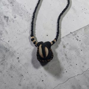 Pode incluir: Um colar com um pingente central feito de um material castanho escuro enrolado em torno de uma concha de búzios cor de creme. O cordão é composto por pequenas contas pretas intercaladas com contas creme perto do pingente.