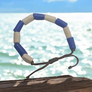 Może przedstawiać: Bransoletka z niebieskich i białych koralików ze sznurkiem do wiązania w kolorze brązowym. Bransoletka wykonana jest z okrągłych, ceramicznych koralików.