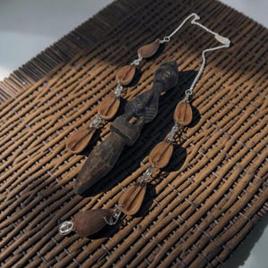 May include: A necklace featuring a central dark brown carved wooden figure with a pointed base, flanked by two silver-toned chains adorned with multiple smooth, light brown, teardrop-shaped wooden beads connected by small silver metal links.