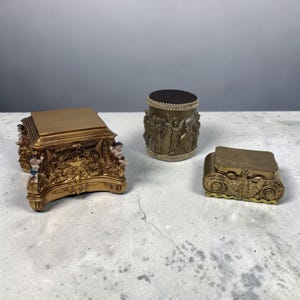 以下が含まれることがあります： 金色のアンティークスタイルの装飾品3点。アイテムには、ケルビムのフィギュアが付いた長方形の箱、レリーフフィギュアが付いた円筒形の容器、装飾的な上部が付いた小さな長方形の箱が含まれます。アイテムは明るい灰色の表面にあります。
