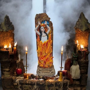 Può includere: Una statuetta di una donna in un abito bianco avvolta dalle fiamme, in piedi su una base di pietra. La statuetta è circondata da candele e altri oggetti rituali, creando un'atmosfera oscura e misteriosa.
