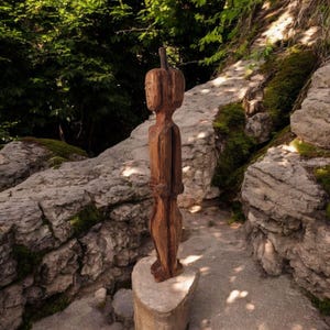 Könnte beinhalten: Eine Holzskulptur einer Person, die auf einem Steinsockel steht. Die Skulptur ist braun und hat ein einfaches, stilisiertes Design. Die Skulptur ist von Felsen und Grün umgeben.