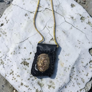 Op de afbeelding: Een zwarte leren hanger met een gebeeldhouwd gezicht, opgehangen aan een gele koord. Het gezicht is lichtbruin met gedetailleerde kenmerken. De hanger ligt op een wit, gebarsten oppervlak.