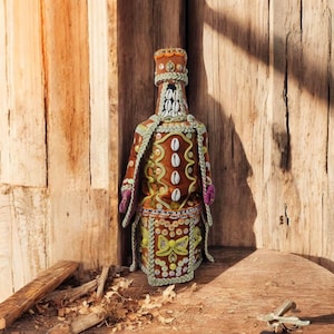 Può includere: Una bottiglia marrone decorata con conchiglie e perline, con una corona e una veste con motivi colorati. La bottiglia è in piedi su una superficie di legno.