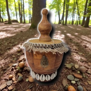 Könnte beinhalten: Ein handgefertigter Holzbehälter mit dekorativer Oberseite und einer mit Muscheln verzierten Basis. Der Behälter hat eine hellbraune Oberfläche, eine Fransenverkleidung und einen Metallakzent. Der Hintergrund ist eine verschwommene Waldszene.