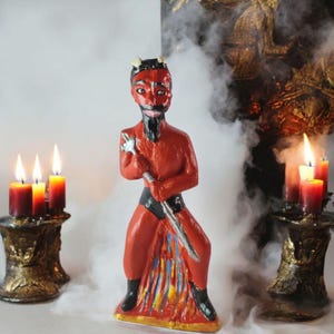 Könnte beinhalten: Eine rote Teufelsfigur mit Hörnern, einem schwarzen Bart und einem schwarzen und silbernen Dreizack. Die Figur steht auf einem Sockel mit darauf gemalten Flammen. Die Figur ist von Kerzen umgeben.