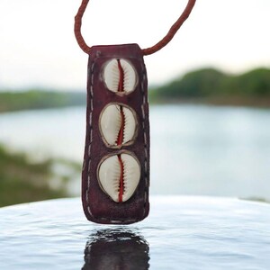 Op de afbeelding: Een bruine leren ketting met drie witte kauri-schelpen eraan bevestigd. De ketting hangt aan een tafel met een wazige achtergrond.