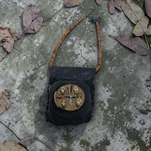 Peut inclure: Petite bourse en cuir noir avec une lanière en cuir marron. Elle présente un emblème circulaire orné de crânes et de croix. Le fond est composé de feuilles sur une surface en béton.