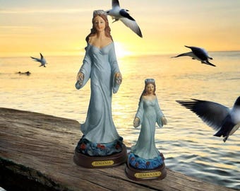 Könnte beinhalten: Zwei blaue und weiße Porzellanstatuen von Yemanja, der Orisha des Ozeans, stehen auf einer Holzoberfläche. Die Statuen tragen Kronen und halten Blumen. Der Hintergrund ist ein Sonnenuntergang über dem Meer.