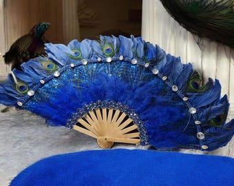 Peut inclure: Un éventail en plumes de paon bleu avec une armature en bois et des strass. L'éventail est fermé et possède une bordure en sequins bleus et argentés.