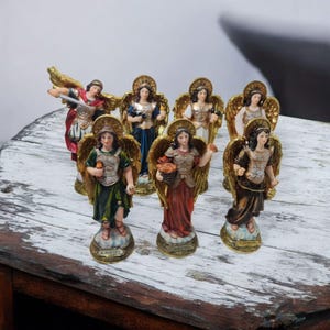 May include: Set of seven small angel figurines, each with gold wings and armour, standing on a white wooden surface. The angels are painted in various colours, including red, blue, green, and brown. Each angel has a name written on the base, including "S. Jophiel", "S. Raphael", and "S. Gabriel".