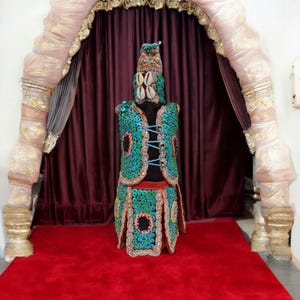 Può includere: Un costume decorativo con un gilet e una gonna blu e verde, adornato con paillettes e conchiglie. Il costume è esposto su un tappeto rosso e una tenda bordeaux.