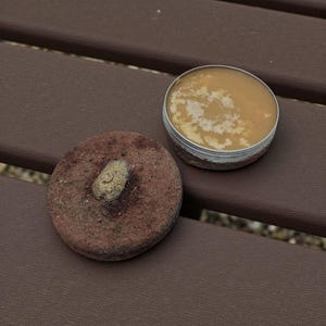 May include: A circular, reddish-brown stone with a lighter, oval-shaped element next to a small, open metal tin. The tin holds a light brown, creamy substance. The items are on a brown surface.