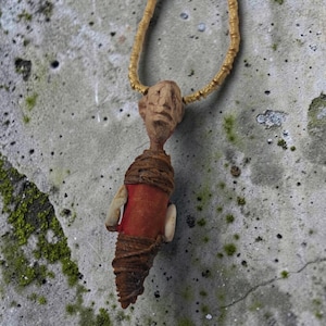 Pode incluir: Um colar com pingente apresentando uma figura humanoide esculpida em tom bege com um torso de tecido vermelho envolto em barbante marrom. A figura está suspensa por uma corrente dourada e possui duas pequenas saliências brancas nas laterais.