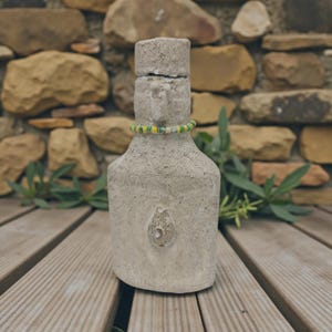 May include: A handmade, pale bottle with a textured finish and a decorative beaded necklace. The bottle has a distinctive shape and a small, textured design on the front. The background features a stone wall and wooden decking.