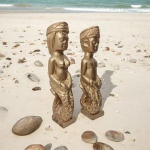 May include: Two gold-colored statues of mermaids with intricate details. The statues are standing on a sandy beach.