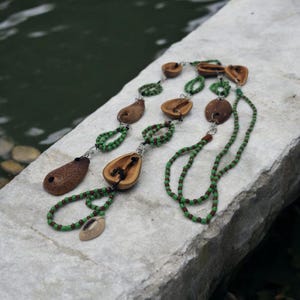 May include: A handmade necklace featuring natural elements. The necklace incorporates brown seed pods and nut shells, connected by strands of green and brown beads. The design includes a variety of shapes and textures, creating a unique, earthy aesthetic.