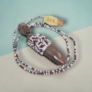 Op de afbeelding: Een bruine houten hangerketting met een gesneden figuur en een kralen lijfje. De ketting heeft een koord van afwisselend witte en kastanjebruine kralen. Een klein label is aan de ketting bevestigd.