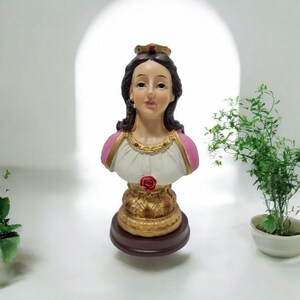Pode incluir: Um busto de resina branca e rosa de uma mulher usando uma coroa e um colar de ouro. O busto está em uma base dourada e marrom.