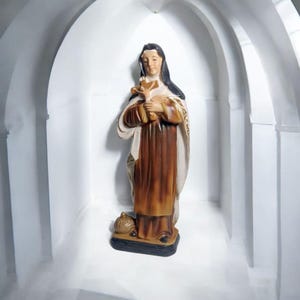 May include: A figurine of a woman in a brown robe and black head covering, holding a cross. The figurine is standing on a black base.