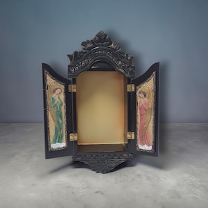 Könnte beinhalten: Ein schwarzer Holzschrank mit zwei Türen, auf denen jeweils ein Engel gemalt ist. Das Innere des Schranks ist hellgoldfarben. Die Türen sind geöffnet und geben den Blick ins Innere frei. Der Schrank hat verzierte Schnitzereien und steht vor einem grauen Hintergrund.