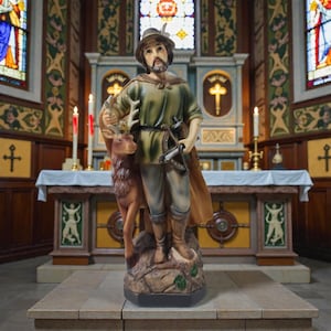 May include: A statue of a man in a green tunic and hat, holding a deer and a crossbow. The statue is in a church setting, with a decorated altar and stained-glass windows in the background.