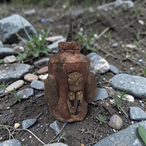 Peut inclure: Un récipient cylindrique marron fait à la main avec une surface texturée. Il présente une figurine dorée sculptée à l'avant et deux éléments latéraux arrondis. Un élément pointu de couleur foncée dépasse du dessus. Le récipient est posé sur un lit de roches et de terre.
