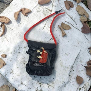 Peut inclure: Une petite pochette noire avec une sangle en cordon rouge. La pochette présente un motif rouge et noir avec des accents dorés. Le fond est une pierre blanche avec des feuilles brunes éparpillées.