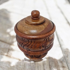 Könnte beinhalten: Ein handgefertigter Holzbehälter mit Deckel. Der Behälter ist dunkelbraun und hat geschnitzte Blumenmuster am Boden. Der Deckel hat einen kleinen Knopf oben. Der Behälter steht auf einer hellen Holzoberfläche. Höhe ca. 10 cm.