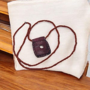 Pode incluir: Uma pequena bolsa de couro bordô escuro com uma abertura circular, suspensa por um cordão marrom a condizer. A bolsa é costurada nas bordas e repousa sobre um tecido branco. O cordão é amarrado para formar um colar.