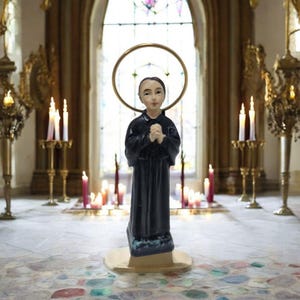 Könnte beinhalten: Eine kleine Statue eines Kindes in einer schwarzen Robe mit ineinander verschränkten Händen zum Gebet. Die Statue steht auf einem goldenen Sockel und hat einen goldenen Heiligenschein über dem Kopf. Die Statue steht vor einem Fenster mit Buntglas und Kerzen.