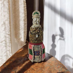 Può includere: Una piccola statuetta decorativa di una persona che indossa una gonna con motivi dorati, verdi, rosa e blu e una corona con conchiglie bianche. La statuetta è realizzata in un materiale marrone e ha una finitura scintillante.