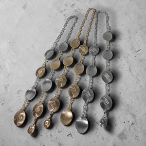 Puede incluir: Cinco cinturones de cadena metálicos en tonos plateados y dorados. Cada cinturón presenta una serie de colgantes circulares y ovalados con diseños geométricos. Los cinturones están dispuestos diagonalmente sobre un fondo gris.