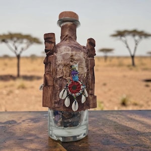Könnte beinhalten: Eine klare Glasflasche, die von geschnitztem braunem Holz mit zwei vertikalen Figuren an den Seiten umhüllt ist. Ein kleiner runder Schmuck mit rotem, blauem und weißem Garn, Perlen und weißen Kaurimuscheln ist in der Mitte des Holzgehäuses befestigt.