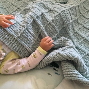 Puede incluir: Una manta de bebé de punto azul claro con un patrón texturizado. La manta está hecha de hilo suave y esponjoso y es perfecta para mantener a un bebé caliente y cómodo.