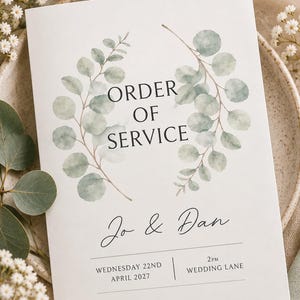 Puede incluir: Una tarjeta blanca de orden de servicio para bodas con un diseño de corona de eucalipto en acuarela. El texto dice: ORDER OF SERVICE, Jo & Dan, WEDNESDAY 22ND APRIL 2027, 2PM, WEDDING LANE. La tarjeta tiene un diseño centrado con fuentes negras.