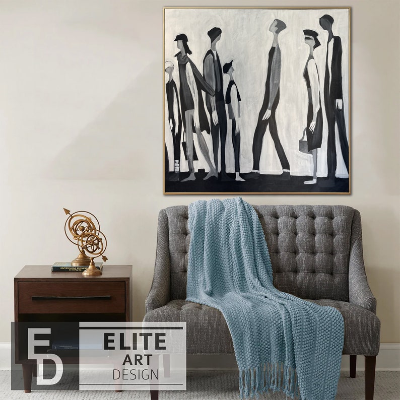 May include: A black and white abstract painting of a group of people walking in a line. The painting is framed in a gold frame and is hanging on a white wall above a gray sofa with a blue throw blanket.