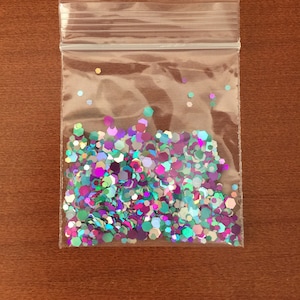 May include: A clear plastic bag filled with iridescent, multi-colored glitter. The glitter is composed of various-sized, hexagonal shapes in shades of purple, teal, and silver. The bag is sealed with a zip-top closure.