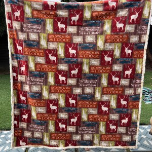 May include: A rectangular blanket with a repeating pattern of deer, trees, and text that reads "WELCOME to the LODGE" and "ADVENTURE BEGINS Here." The blanket has a white sherpa trim and is in shades of red, brown, and green.