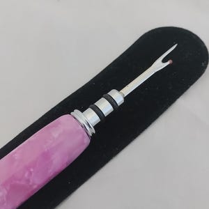 May include: A seam ripper featuring a polished silver metal tip with a small red safety ball, a silver ferrule with two black rubber rings, and a cylindrical handle with a marbled pink and white acrylic finish, resting on a black velvet sleeve.