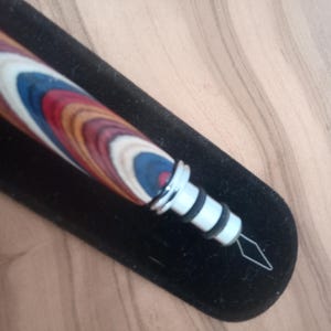 May include: A wooden pen with a multi-colored design of red, blue, white, and brown. The pen has a silver metal tip and is resting on a black velvet surface. The pen is a writing instrument.
