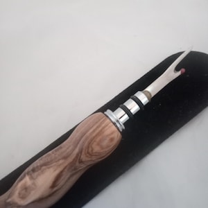 May include: A seam ripper with a wooden handle and a silver metal blade. The handle has a light brown wood grain pattern. The blade has a curved tip with a small red ball at the end. The seam ripper is resting on a black surface.