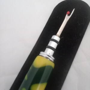 May include: A seam ripper with a silver blade and a handle with a green and yellow swirl pattern. The seam ripper has a red ball at the tip and is set against a black background.