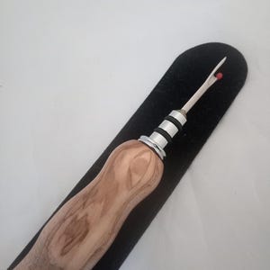 May include: A seam ripper with a light brown wooden handle and a silver metal blade. The blade has a small red ball at the tip. The seam ripper is resting on a black felt surface.