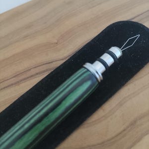 May include: A needle threader featuring a cylindrical handle with a green and black striped pattern. The handle is accented with silver-toned metal rings and a black rubber grip section. A thin, diamond-shaped wire loop extends from the tip.