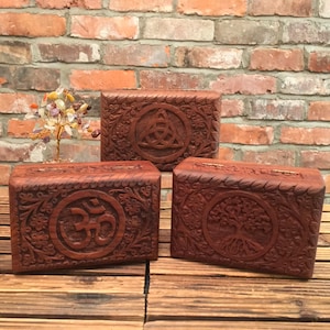 May include: Three wooden boxes with intricate carvings. The boxes feature a Celtic knot, an Om symbol, and a tree of life design. The boxes are made of dark brown wood and have a polished finish.