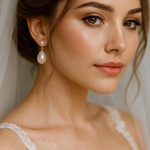 May include: A woman wearing pearl drop earrings. The earrings have a small round stud and a large teardrop-shaped pearl. She is wearing a white lace dress and a sheer veil. Her makeup includes blush and lipstick.